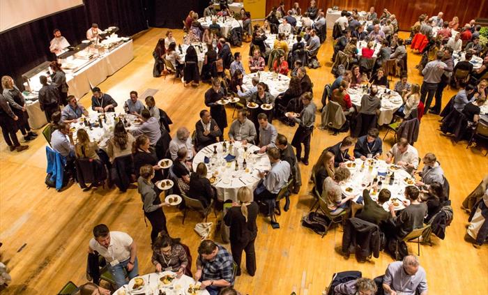 A busy conference dinner, guests being served by staff to round tables.